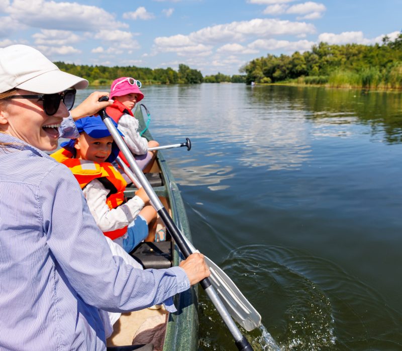 Woman two children enjoy having fun paddling canoe rent across lake river water wearing bright orange life vests hot sunny summer day. Healthy recreational activity lifestyle vacation trip journey.